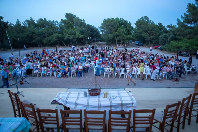 Το κοινό σε πλαστικές καρέκλες, κάτω από πεύκα, μπροστά σε υπαίθρια σκηνή με τραπέζι και ψάθινες καρέκλες