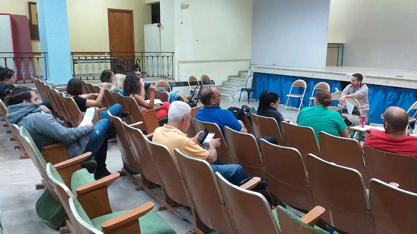 Ο νέος σκηνοθέτης μιλά στην ομάδα από το μπροστινό μέρος της αίθουσας, ενώ τα μέλη κάθονται στις σειρές των καθισμάτων